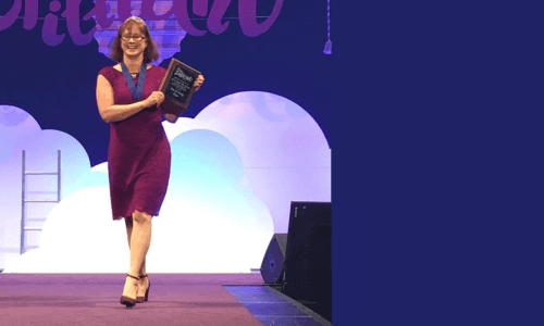 photo of Becky Dean in an eggplant knee-length lace dress walking on stage with an Usborne Books & More award plaque.