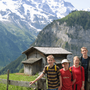 photo of Tom & Becky Dean's 4 children standing in a green grass meadow with a Switzerland snow covered mountain vista scene behind in the distance.