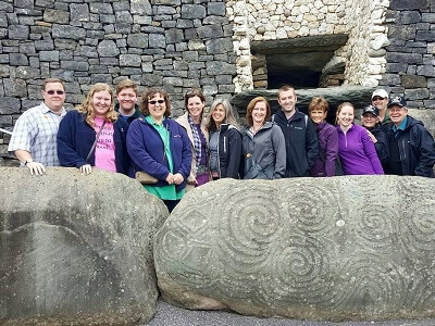 Tom & Becky Dean and several Book Nation team members at the Newgrange tomb in Ireland - standing behind the very large entrance kerbstone with megalithic art engraved on it and the tomb in the background.