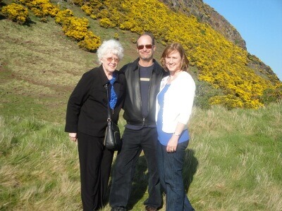 Tom & Becky Dean with Becky's mom in Scotland - a hill of green and yellow flower beyond
