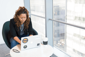 brown skinned woman with brown dreadlocks seated at a table next to a window looking at a computer